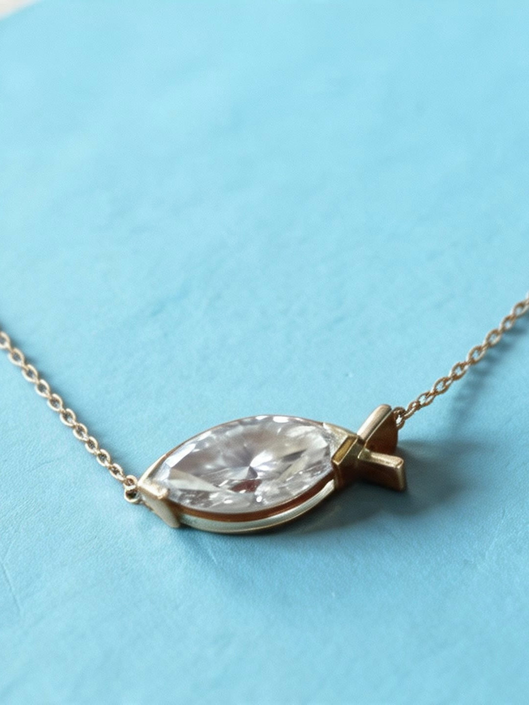 A gold necklace with a east west marquise diamond pendant and a small cross-shaped accent, displayed on a textured light blue surface.