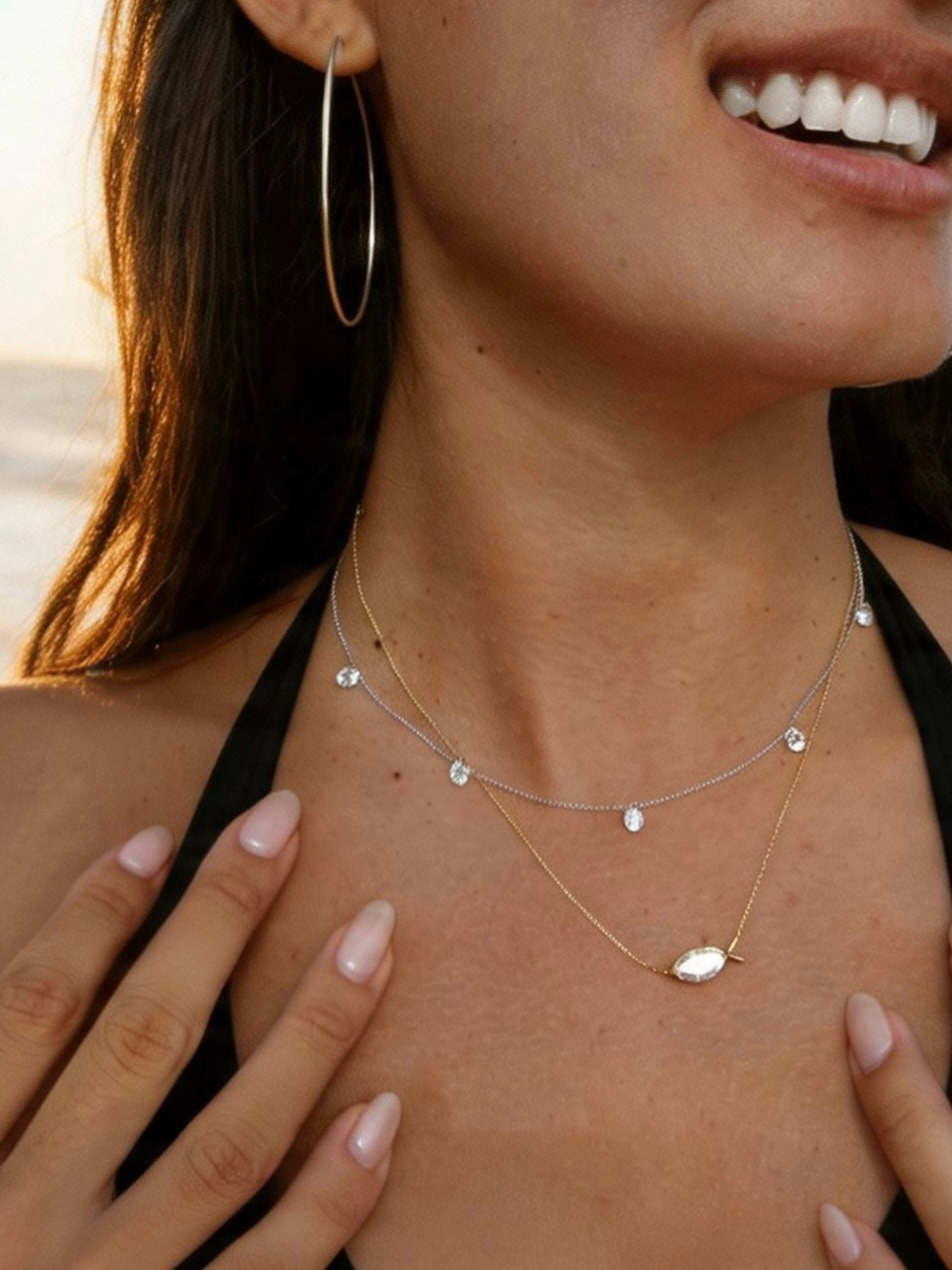 A close-up lifestyle shot of a woman wearing layered necklaces. The necklaces include a silver chain with 5 small diamonds and a gold chain with a marquise diamond pendant in the shape of a fish.